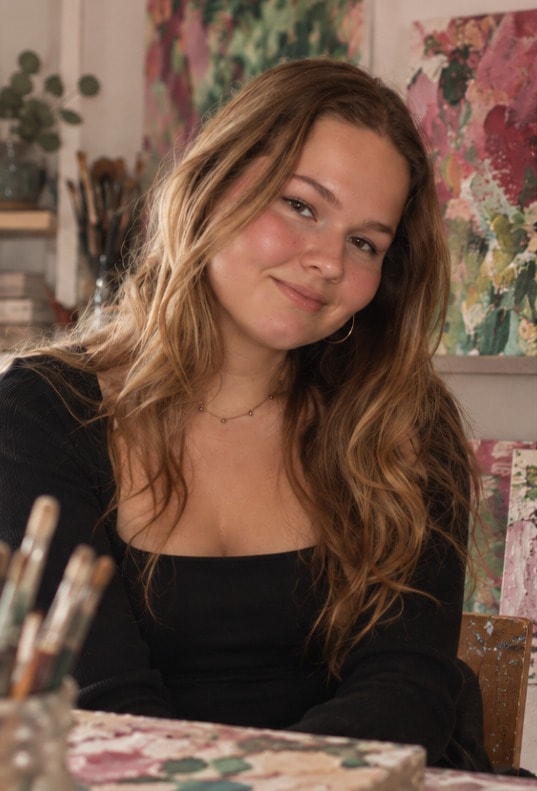 Daphne Merel, smiling, at a table in her studio, with brushes in a jar and colourful paintings behind her.