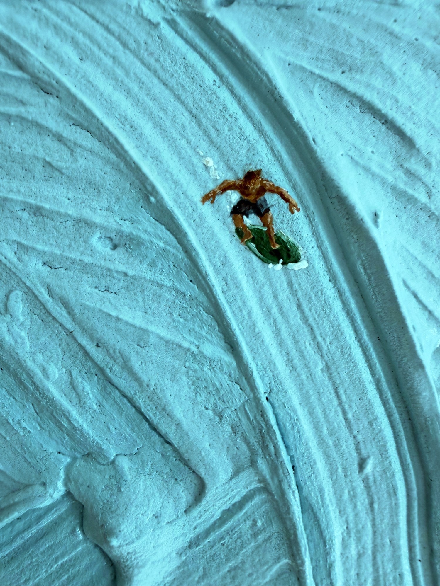 Detail van een kleine surfer op een groene plank die over een gebogen rug van turquoise verf glijdt.