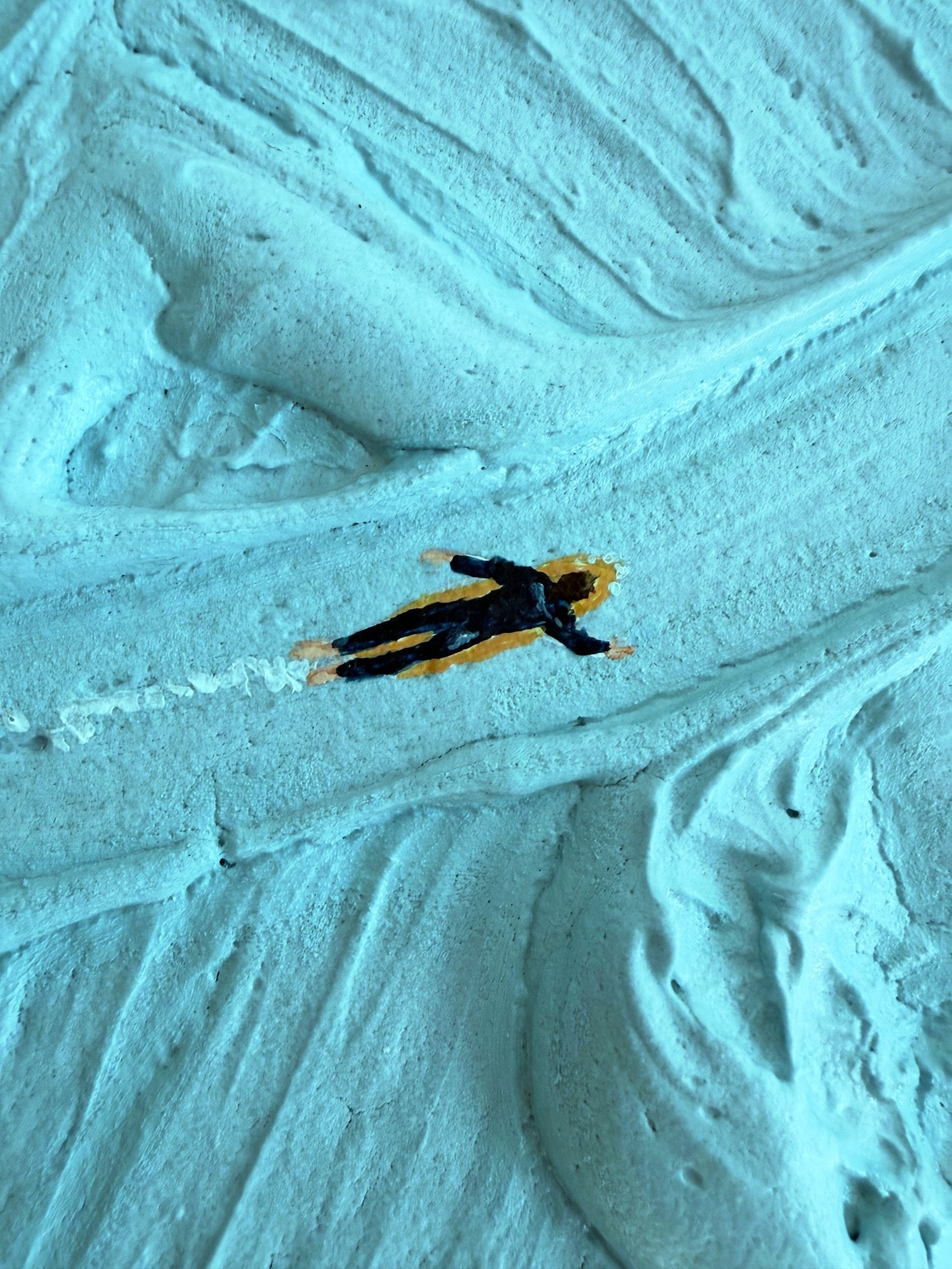 Detail van een klein figuurtje in een zwart wetsuit op een oranje plank, met een wit kielzog erachter.